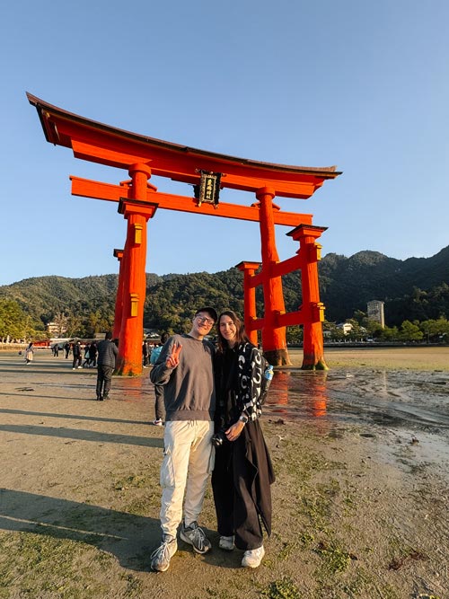 nele and richard standing on wet sand in front of the grand torii gate of itsukushima shrine on miyajima island