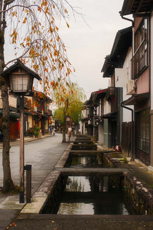 wooden lantern next to a small canal with koi swimming in it, traditional japanese houses on the side and some small willow trees